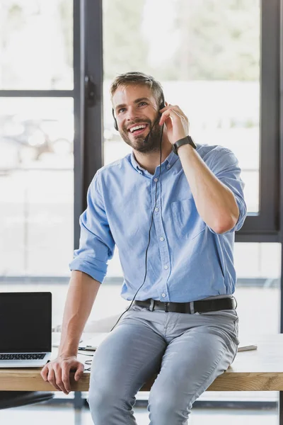Handsome smiling young call center operator in headset sitting on table in office — Stock Photo