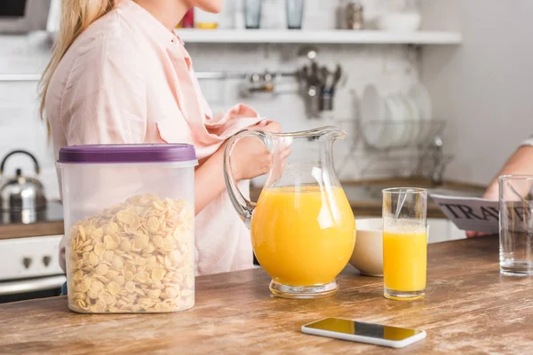 Обрізане зображення дівчини, що застібає блузку біля столу з апельсиновим соком та кукурудзяними пластівцями на сніданок на кухні — стокове фото