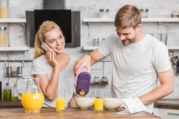 Fidanzato versando cornflakes nel piatto durante la colazione in cucina, fidanzata che parla con lo smartphone — Foto stock