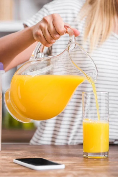 Imagen recortada de la mujer vertiendo jugo de naranja en vidrio en la cocina - foto de stock