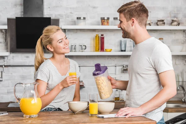 Boyfriend holding container of cornflakes and looking at girlfriend during breakfast in kitchen — Stock Photo
