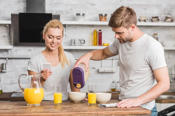 Freundin mit Smartphone und Freund gießen beim Frühstück in Küche Cornflakes in Teller — Stockfoto