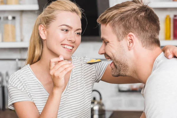 Retrato de sorrindo namorada alimentando namorado com flocos de milho na cozinha — Fotografia de Stock