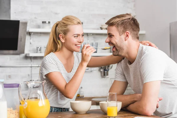 Fidanzata sorridente alimentazione fidanzato con cornflakes in cucina — Foto stock