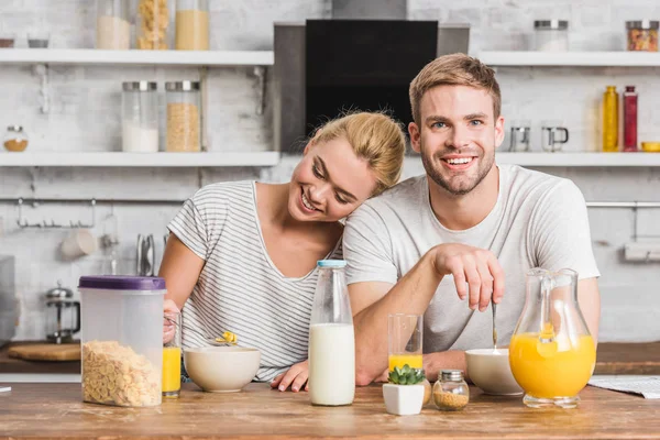 Casal feliz sentado à mesa com leite, suco e flocos de milho na cozinha — Fotografia de Stock