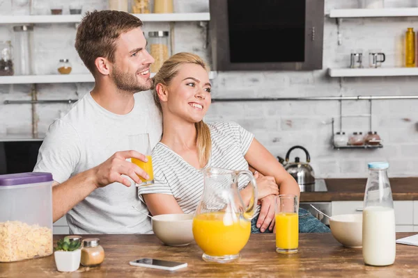 Namorado feliz abraçando namorada durante o café da manhã e segurando vidro de suco na cozinha — Fotografia de Stock