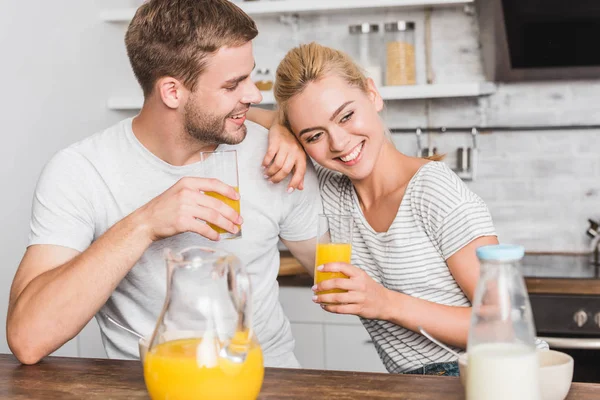 Casal feliz segurando copos de suco de laranja e abraçando na cozinha — Fotografia de Stock