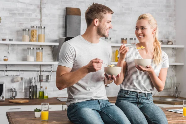 Alegre pareja sosteniendo platos y cucharas con copos de maíz y mirándose en la cocina - foto de stock