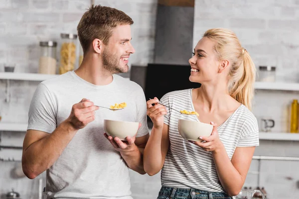 Feliz pareja sosteniendo platos y cucharas con hojuelas de maíz y mirándose en la cocina - foto de stock