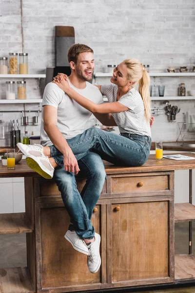 Glückliches heterosexuelles Paar, das sich umarmt und auf dem Küchentisch sitzt — Stockfoto