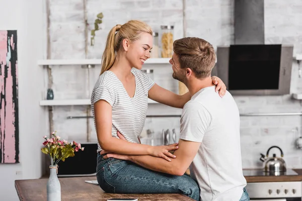 Vista laterale della fidanzata felice seduta sul bancone della cucina e guardando il fidanzato — Foto stock