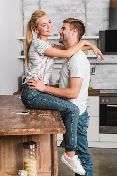 Petite amie heureuse assise sur le comptoir de la cuisine et câlin petit ami — Photo de stock