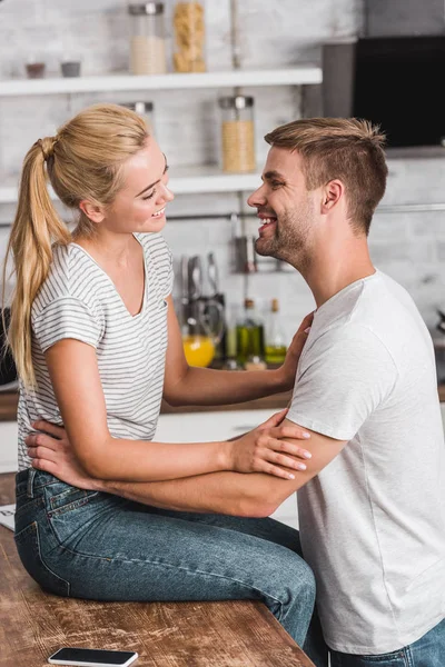 Feliz namorada sentada no balcão da cozinha e olhando para o namorado — Fotografia de Stock