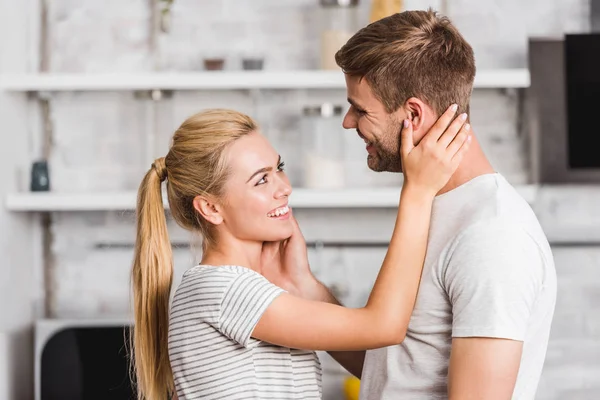 Seitenansicht eines lächelnden Paares, das sich in der Küche umarmt und Gesichter berührt — Stockfoto