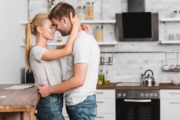 Vue latérale du couple câlin dans la cuisine et penché sur le comptoir de la cuisine — Photo de stock