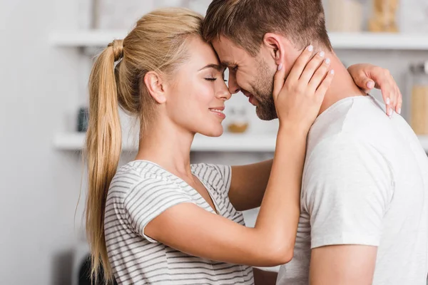 Vista lateral de pareja cariñosa abrazándose en cocina y tocando con frentes - foto de stock