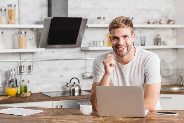 Sonriente hombre guapo trabajando con el ordenador portátil en la cocina y mirando a la cámara - foto de stock
