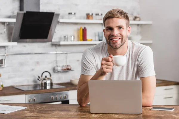 Schöner lächelnder Mann mit Tasse Kaffee und Blick in die Kamera in der Küche — Stockfoto