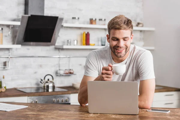 Bell'uomo sorridente che tiene la tazza di caffè e guarda il computer portatile in cucina — Foto stock