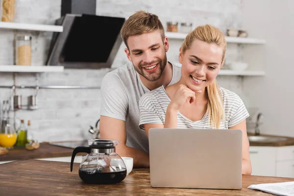 Happy boyfriend hugging girlfriend in kitchen and they looking at laptop — Stock Photo