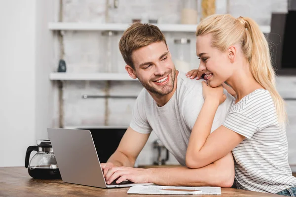 Smiling girlfriend hugging handsome boyfriend in kitchen while he working with laptop — Stock Photo