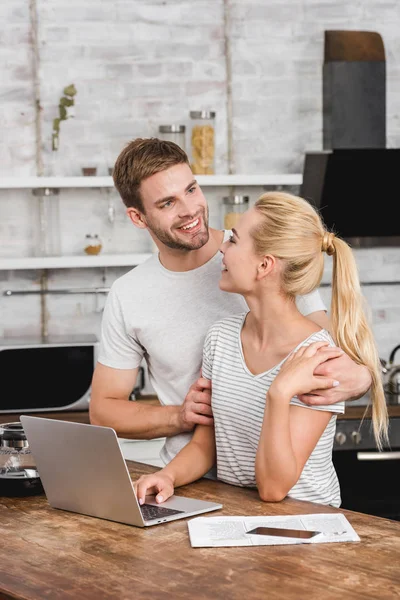 Novio sonriente abrazando novia en la cocina mientras ella trabaja con el ordenador portátil - foto de stock