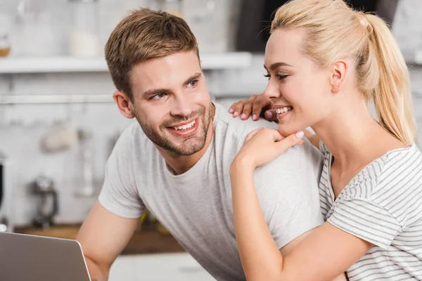 Novia sonriente abrazando novio en la cocina mientras trabaja con el ordenador portátil — Stock Photo