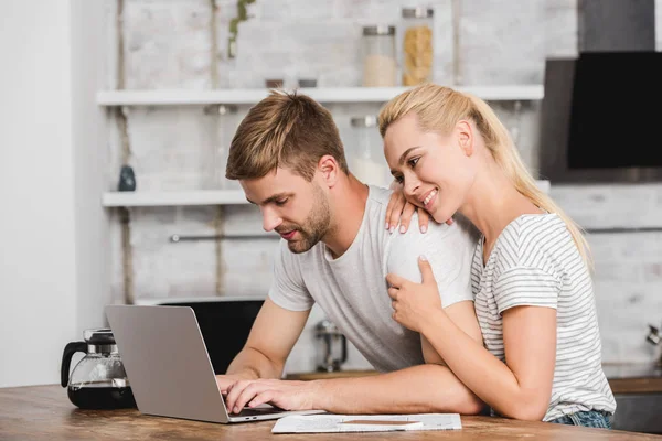 Cheerful girlfriend hugging boyfriend in kitchen and he working with laptop — Stock Photo