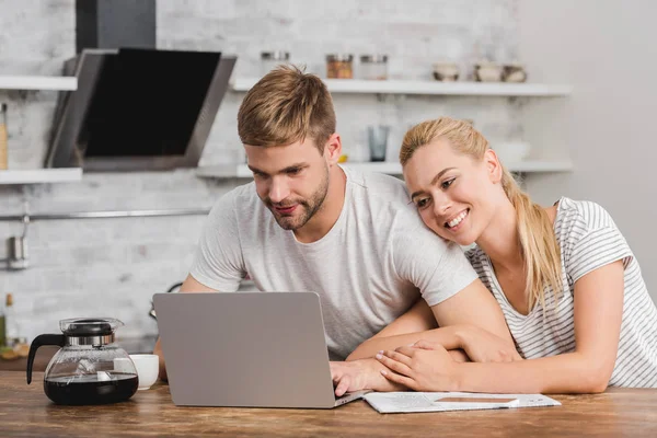 Smiling girlfriend hugging boyfriend in kitchen and he using laptop — Stock Photo