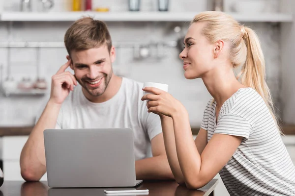 Smiling couple sitting in kitchen with laptop and cup of coffee — Stock Photo
