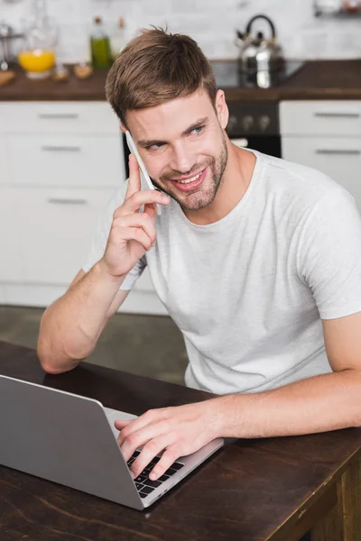 Smiling young man talking by smartphone and using laptop at home — Stock Photo