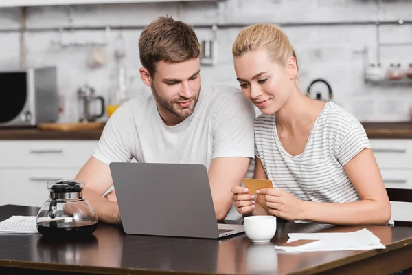 Sonriente joven pareja sosteniendo tarjeta de visita y el uso de ordenador portátil juntos en la mesa de la cocina - foto de stock