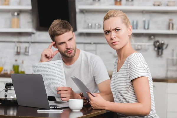 Frowning young woman holding papers and looking at camera while using laptop with boyfriend at home — Stock Photo