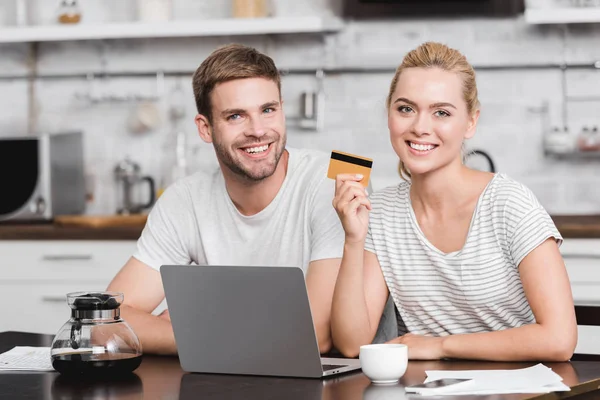 Felice giovane coppia con carta di credito utilizzando il computer portatile e sorridendo alla fotocamera a casa — Foto stock