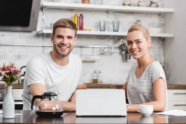 Feliz joven pareja usando el ordenador portátil y sonriendo a la cámara en la cocina - foto de stock