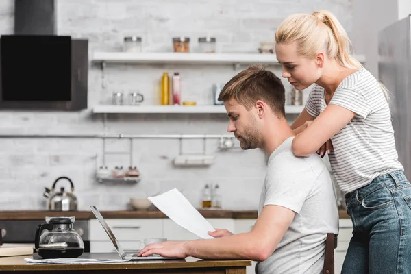 Vista lateral do jovem casal olhando para folha de papel e usando laptop juntos em casa — Fotografia de Stock