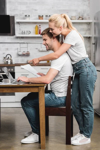 Feliz joven mujer abrazando novio sonriente y señalando con el dedo a la computadora portátil - foto de stock