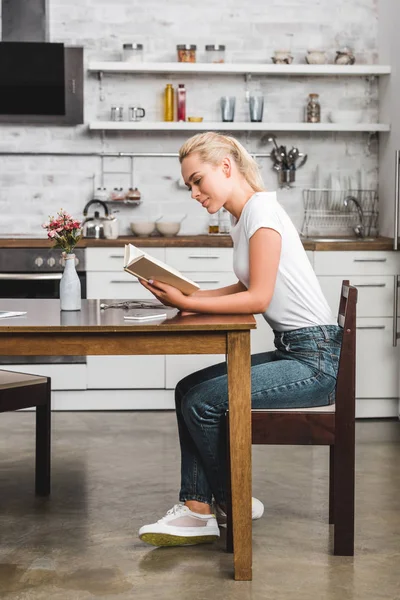 Vue latérale d'une belle jeune femme lisant un livre assis à table dans la cuisine — Photo de stock