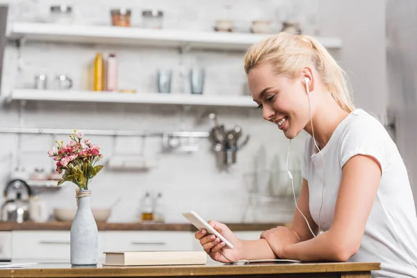 Seitenansicht einer lächelnden jungen Frau mit Kopfhörern, die ihr Smartphone zu Hause benutzt — Stockfoto