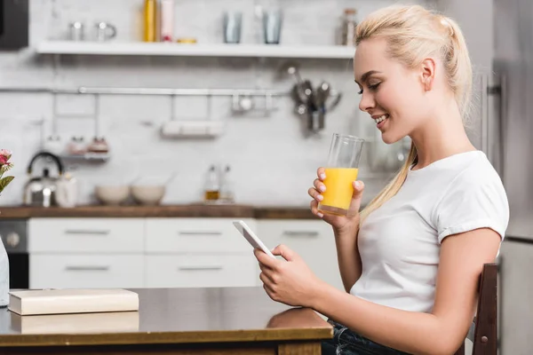 Vista laterale di sorridere giovane donna bere succo di frutta fresco e utilizzando smartphone a casa — Foto stock