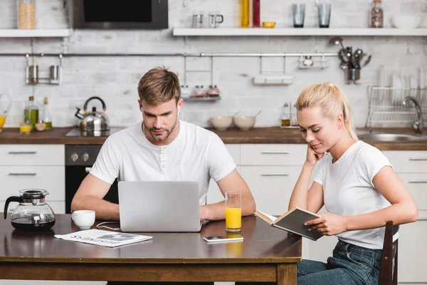 Jovem homem usando laptop e mulher sorridente leitura livro enquanto sentados juntos na mesa da cozinha — Fotografia de Stock