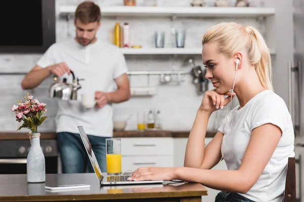 Attractive young woman in earphones using laptop while boyfriend holding cup and kettle in kitchen — Stock Photo