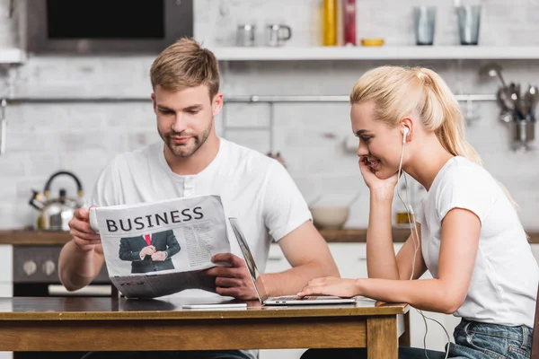 Joven guapo leyendo periódico de negocios y novia sonriente en auriculares usando el ordenador portátil en casa - foto de stock