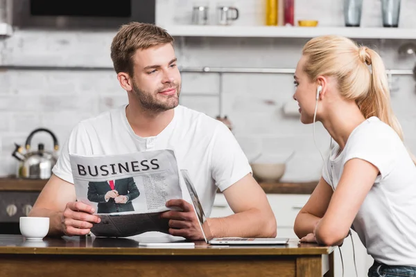 Smiling young man reading business newspaper and looking at happy girlfriend in earphones while sitting at kitchen table — Stock Photo