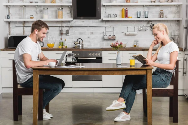 Seitenansicht eines glücklichen jungen Paares, das beim Frühstück am Küchentisch Buch liest und Laptop benutzt — Stockfoto