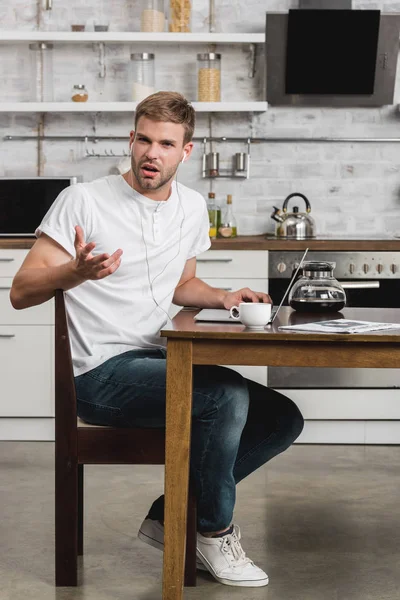 Емоційний молодий чоловік в навушниках, використовуючи ноутбук, сидячи за столом на кухні — стокове фото