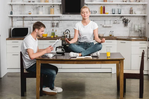 Young man in earphones reading newspaper while beautiful girlfriend meditating on kitchen table — Stock Photo