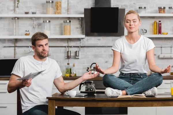 Jovem segurando jornal e olhando para a câmera enquanto bela namorada meditando na mesa da cozinha — Fotografia de Stock