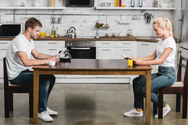 Side view of young couple reading book and newspaper while drinking coffee and juice at morning — Stock Photo