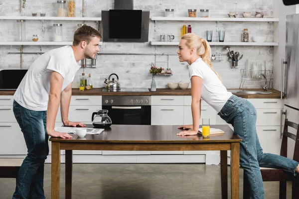 Вид сбоку страстной молодой пары, смотрящей друг на друга, читающей и пьющей кофе с соком на кухне — стоковое фото